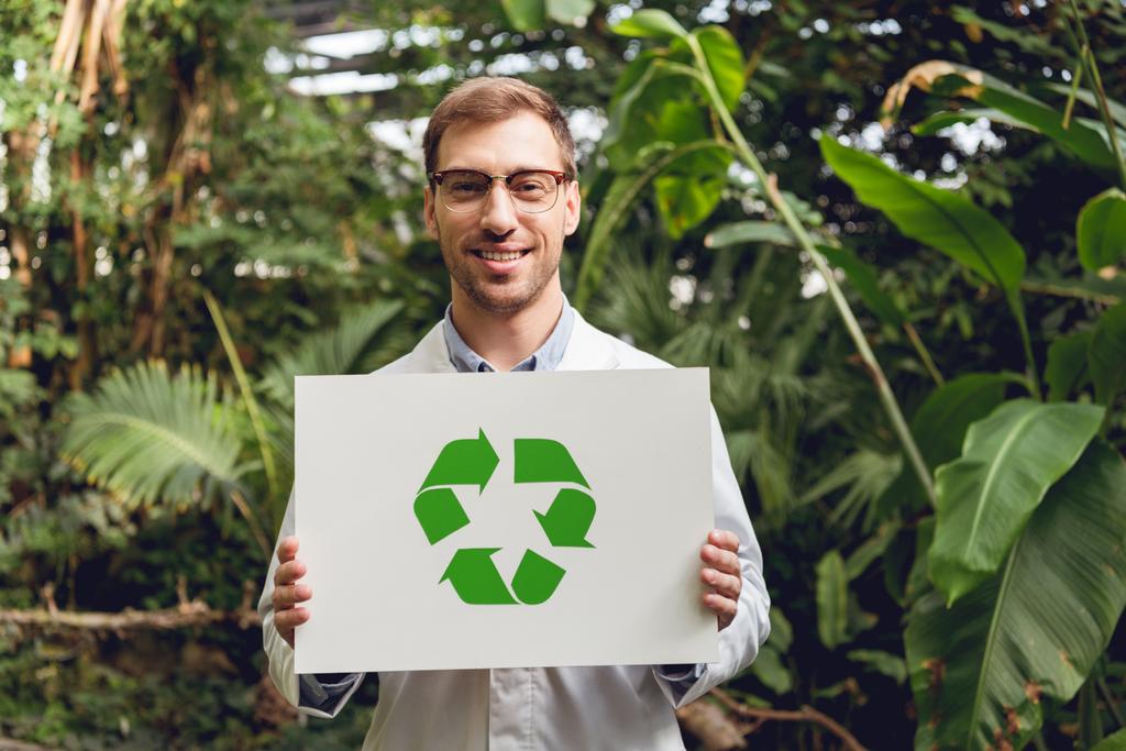 cientista bonito sorridente em casaco branco e óculos cartão de segurança com sinal de reciclagem verde em laranjaria
 - Foto, Imagem
