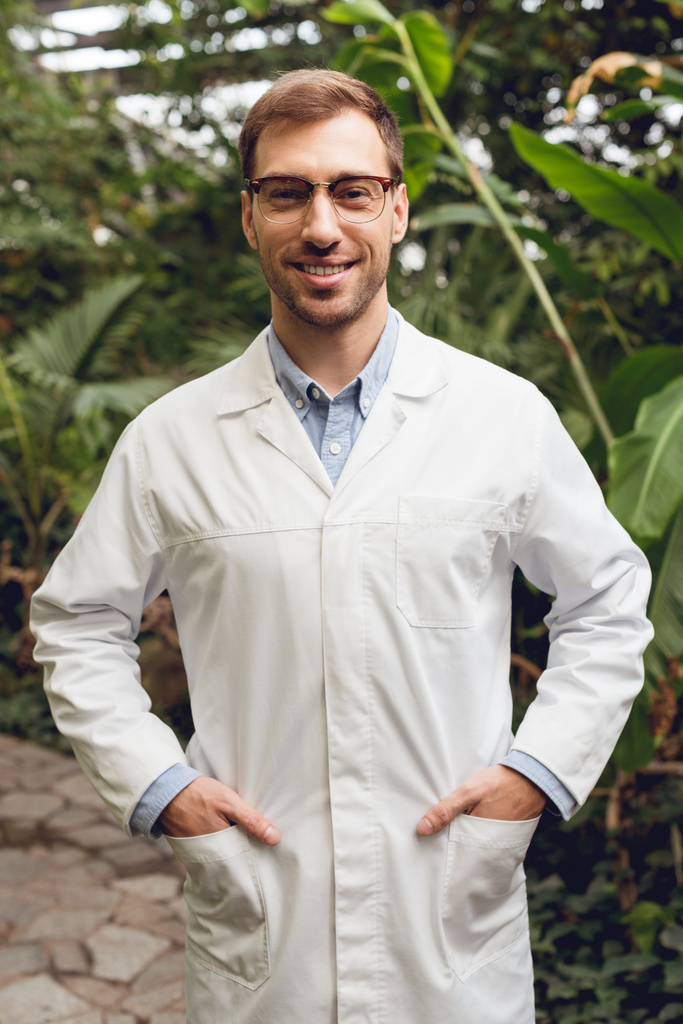 glimlachend knappe wetenschapper in witte vacht en glazen staande met handen in zakken in Oranjerie - Foto, afbeelding