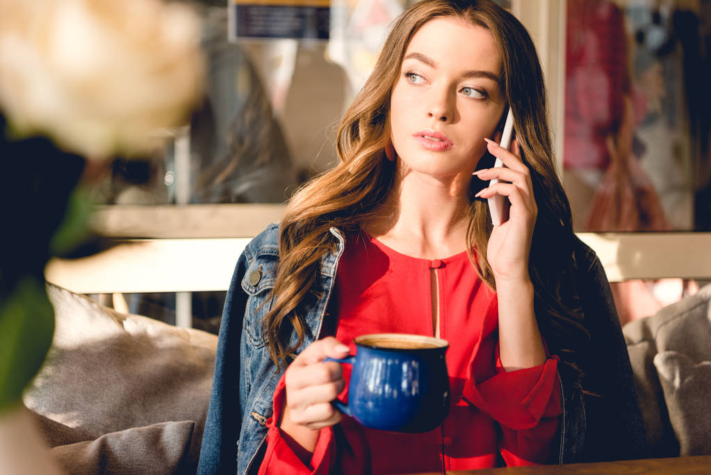 コーヒーのカップを保持し、カフェでスマートフォンで話す魅力的な若い女性  - 写真・画像