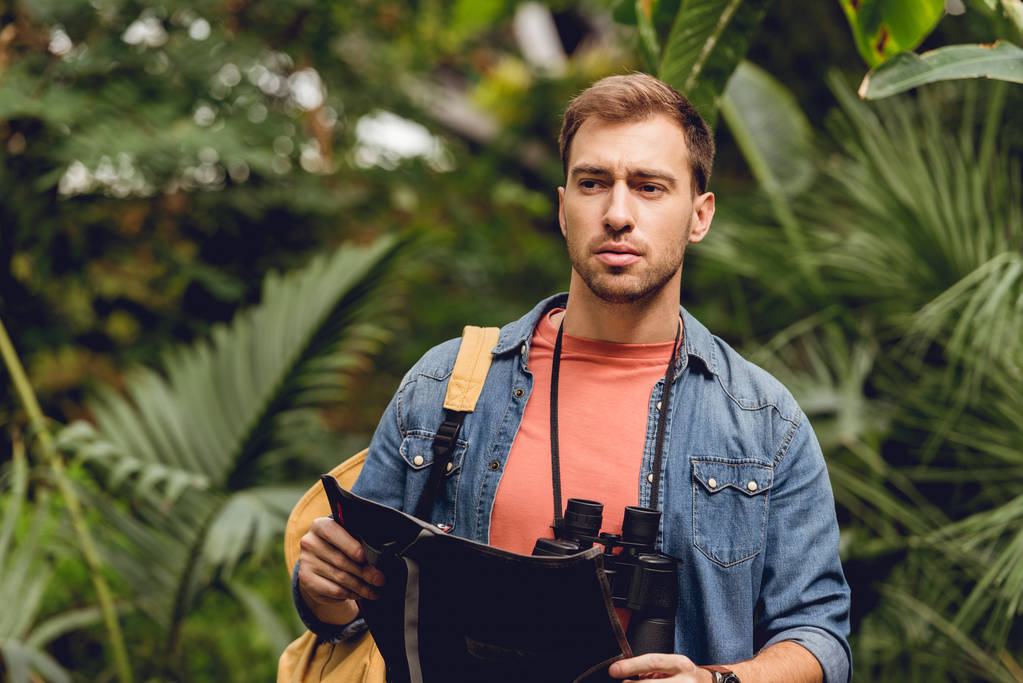 handsome tense traveler with binoculars and backpack holding map in green tropical forest - Photo, Image