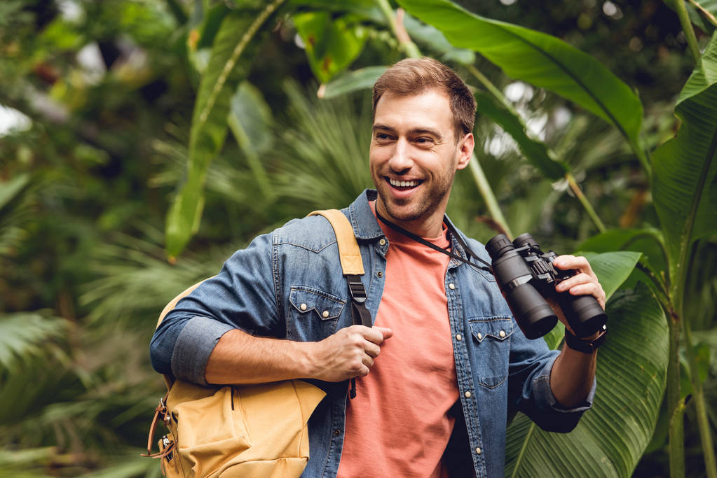 guapo viajero sonriente con prismáticos y mochila en bosque tropical verde
 - Foto, Imagen