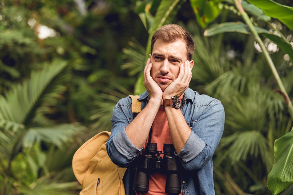 handsome sad traveler with binoculars and backpack in green tropical forest - Photo, Image