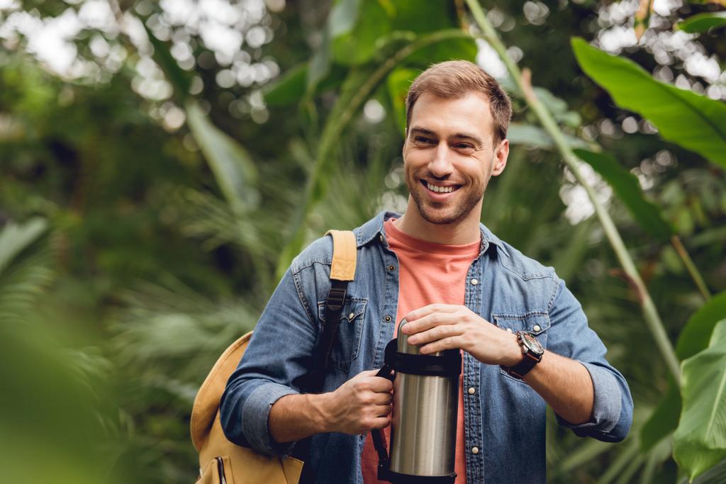 селективный фокус улыбающегося путешественника с рюкзаком открытия термоса в тропическом лесу
 - Фото, изображение