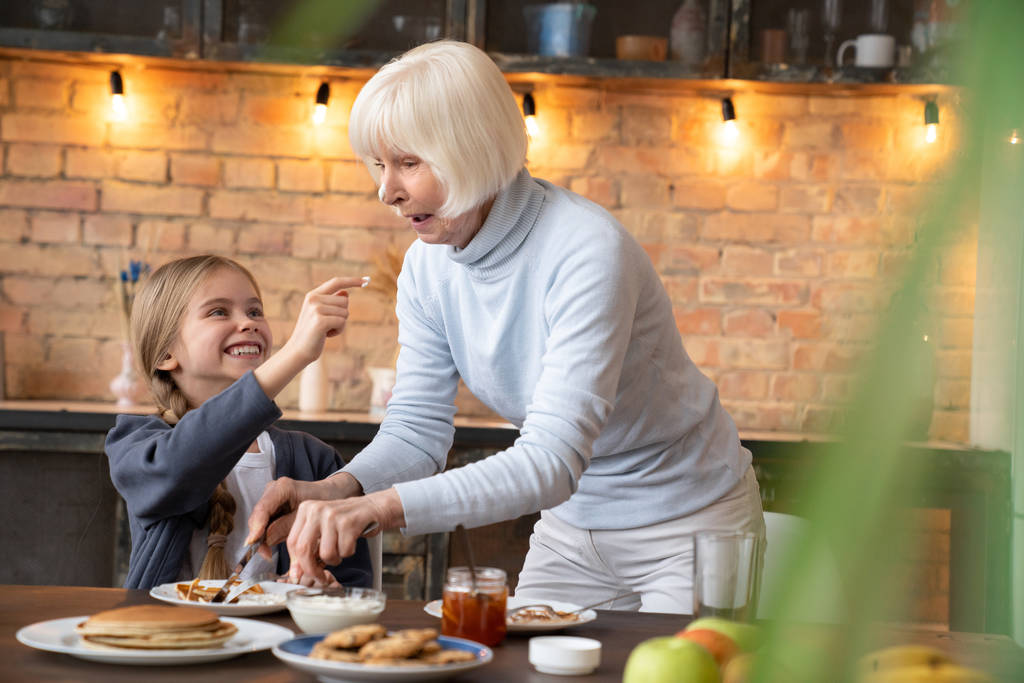 Горизонтальний знімок щасливої маленької дівчинки розважається зі своєю бабусею на кухні біля столу під час сніданку
 - Фото, зображення