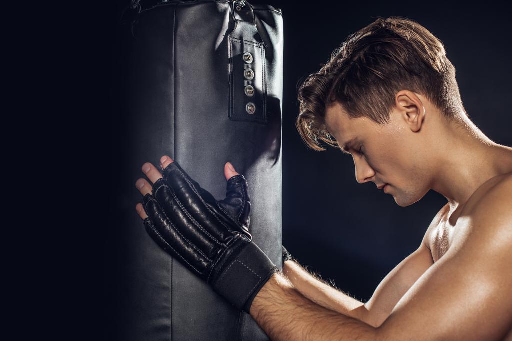 Boční pohled na boxera držící boxovací vak se zavřenýma očima na černém - Fotografie, Obrázek