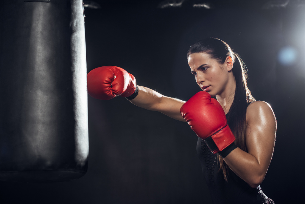 Female boxer in red boxing gloves training with punching bag on black - Photo, Image