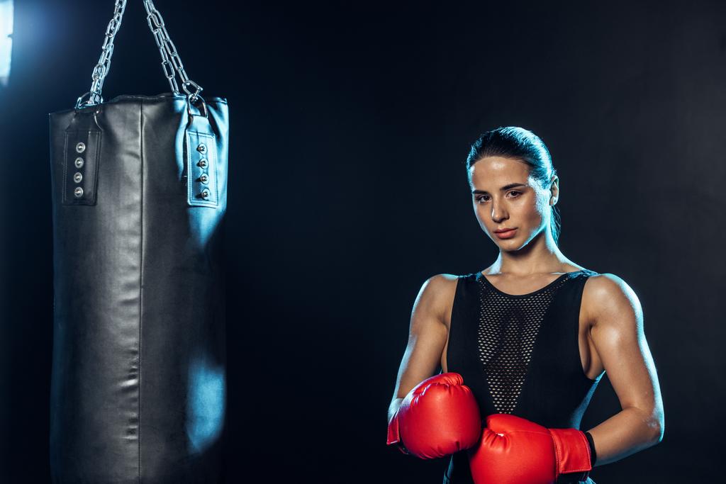 Strong boxer in red gloves standing near punching bag and looking at camera on black - Photo, Image