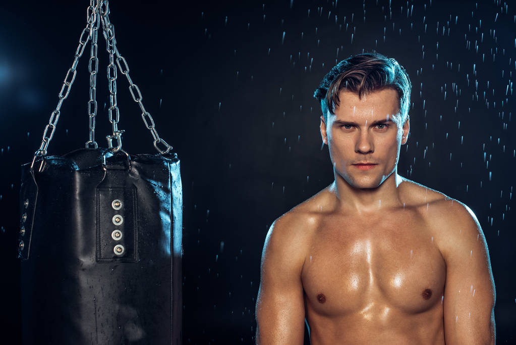Frontansicht eines Boxers, der in der Nähe eines Boxsacks unter Wassertropfen auf schwarz steht - Foto, Bild