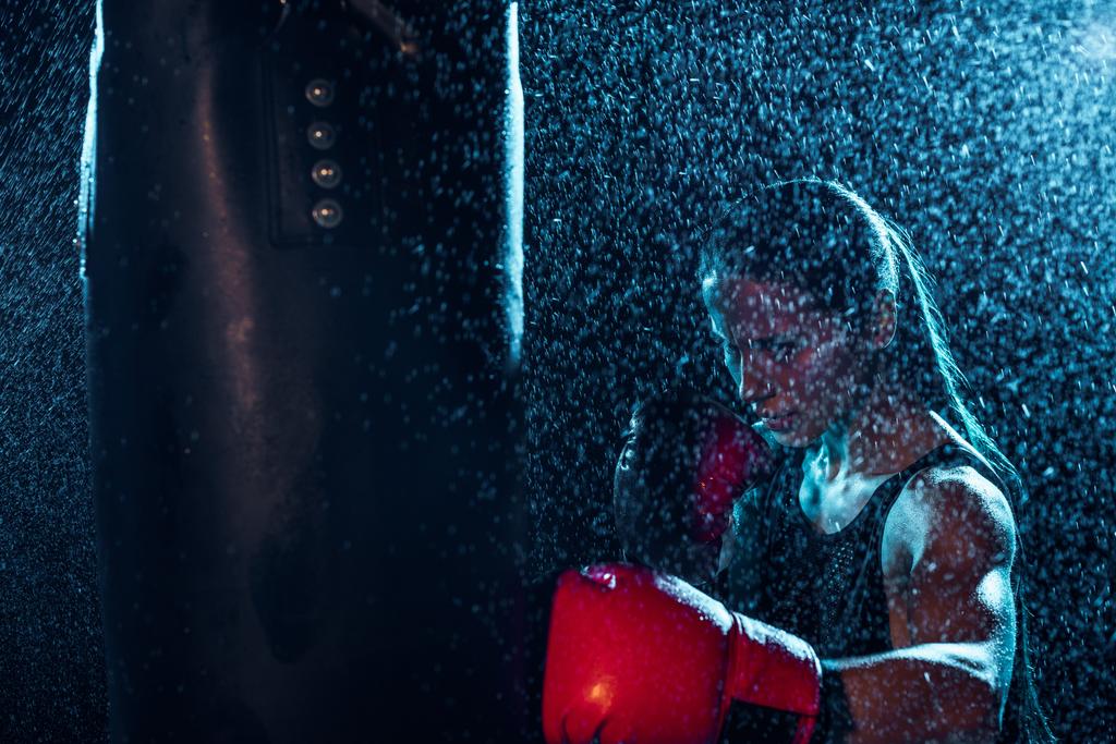 Boxeador fuerte en guantes de boxeo rojos de entrenamiento bajo gotas de agua en negro
 - Foto, imagen