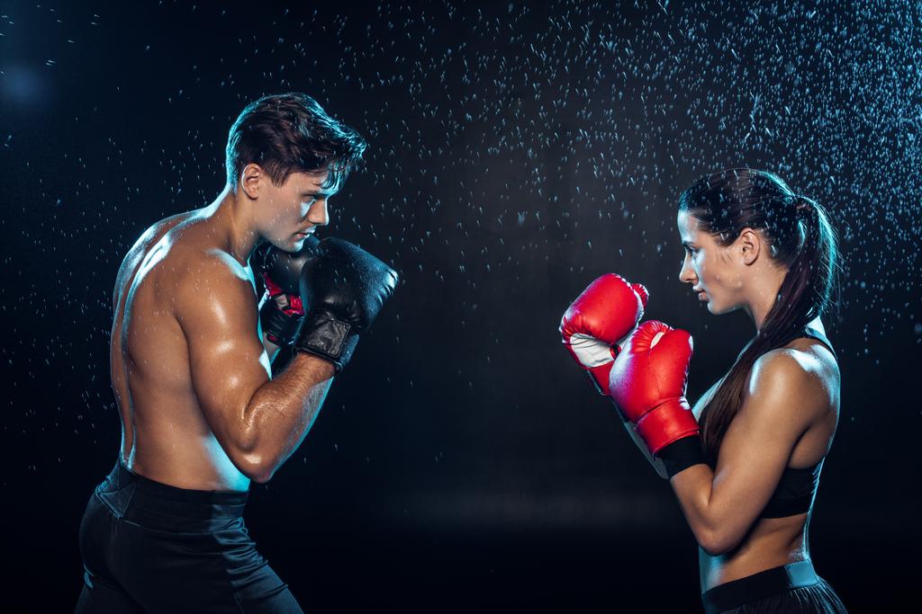 Seitenansicht von zwei Boxern in Boxhandschuhen, die zusammen unter Wassertropfen auf schwarz trainieren - Foto, Bild