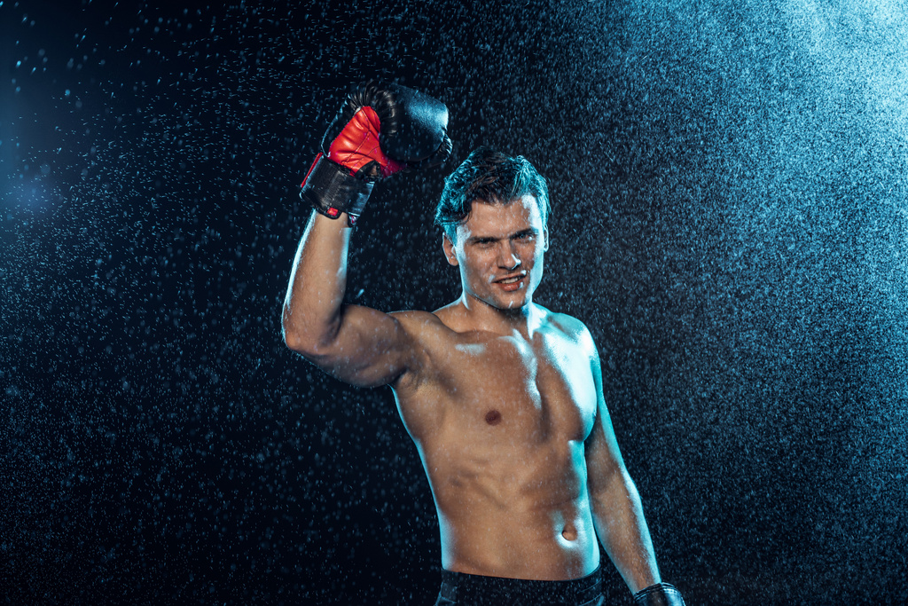 Glimlachende Boxer staande met de hand onder water druppels op zwart - Foto, afbeelding
