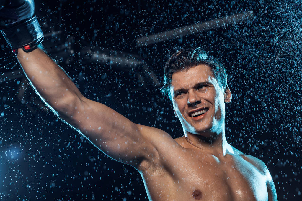 Lächelnder Boxer steht mit erhobener Hand unter Wassertropfen auf schwarz - Foto, Bild