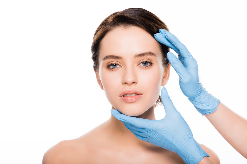 обрезанный вид пластического хирурга в голубых латексных перчатках трогательное лицо красивой женщины изолированы на белом
 - Фото, изображение