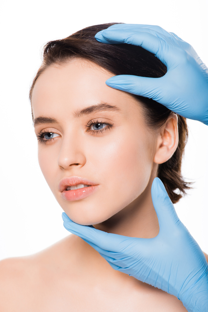 обрізаний вид пластичного хірурга в синіх латексних рукавичках, що торкаються обличчя красивої жінки, ізольованої на білому
 - Фото, зображення