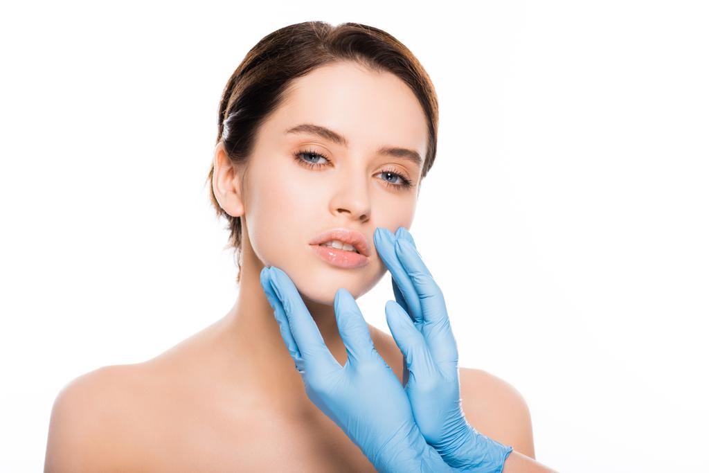 περικομμένη όψη του πλαστικού χειρουργού σε γάντια λατέξ αγγίζοντας το πρόσωπο της μελαχρινή γυναίκα απομονωθεί σε λευκό - Φωτογραφία, εικόνα