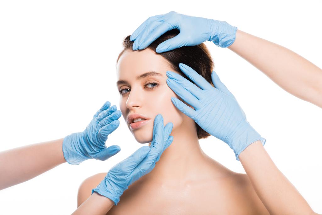 bijgesneden weergave van plastische chirurgen in blauwe latex handschoenen raken gezicht van naakte meisje te kijken naar de camera geïsoleerd op wit - Foto, afbeelding