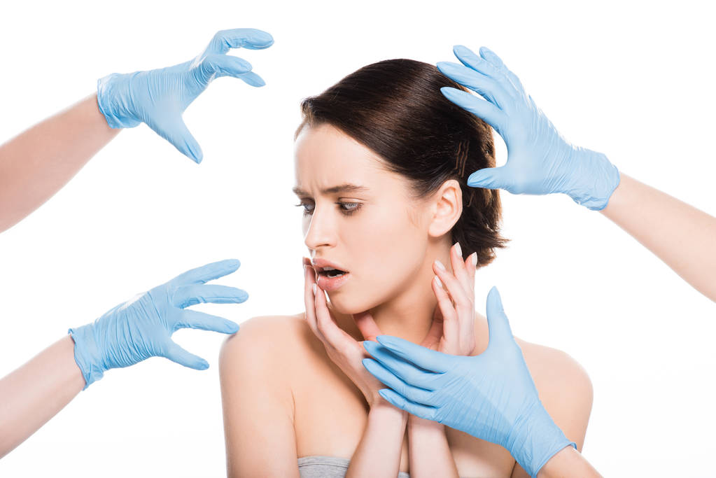 abgeschnittene Ansicht von plastischen Chirurgen in blauen Latex-Handschuhen in der Nähe des Gesichts eines schockierten Mädchens isoliert auf weiß - Foto, Bild