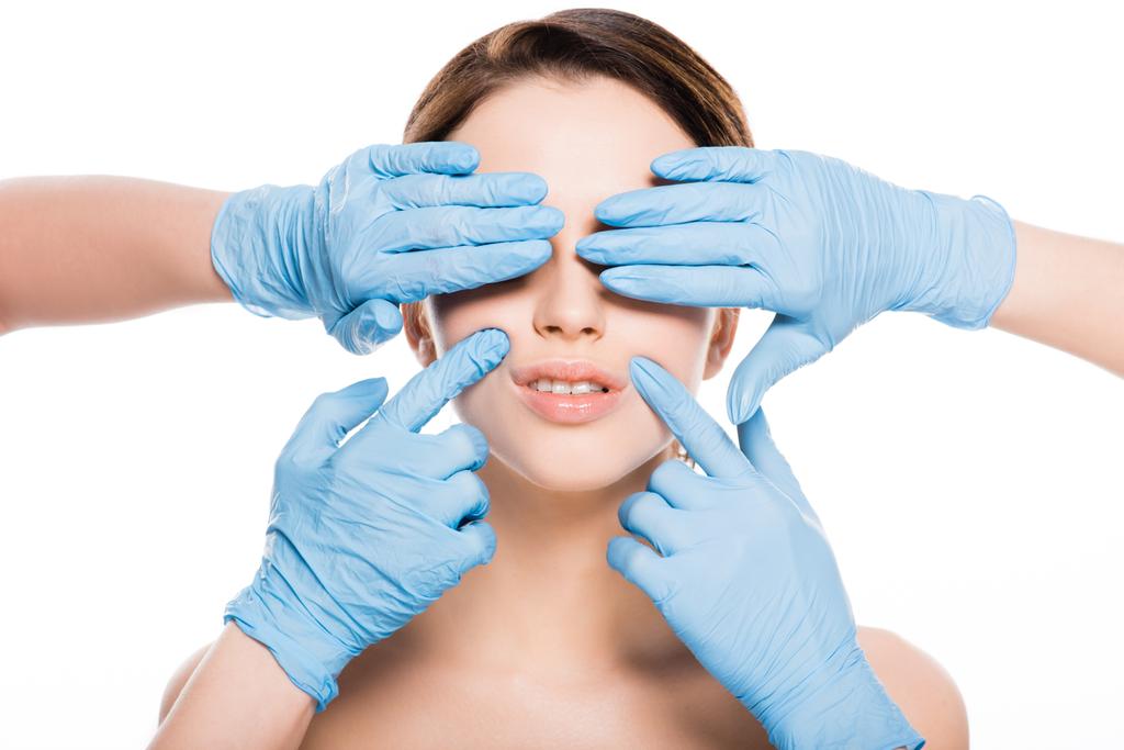 Ausgeschnittene Ansicht von plastischen Chirurgen in blauen Latex-Handschuhen, die Augen bedecken und das Gesicht eines Mädchens berühren, das isoliert auf weiß ist - Foto, Bild