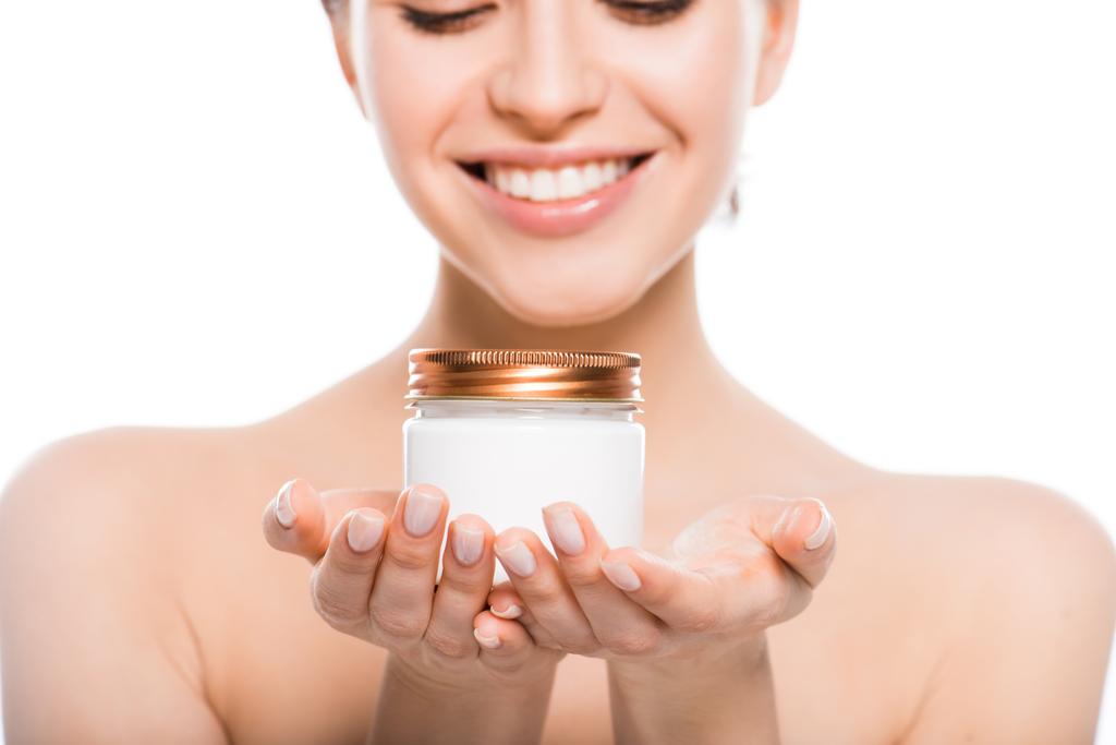 bijgesneden weergave van naakte jonge vrouw Holding container met gezichtscrème en glimlachend geïsoleerd op wit  - Foto, afbeelding