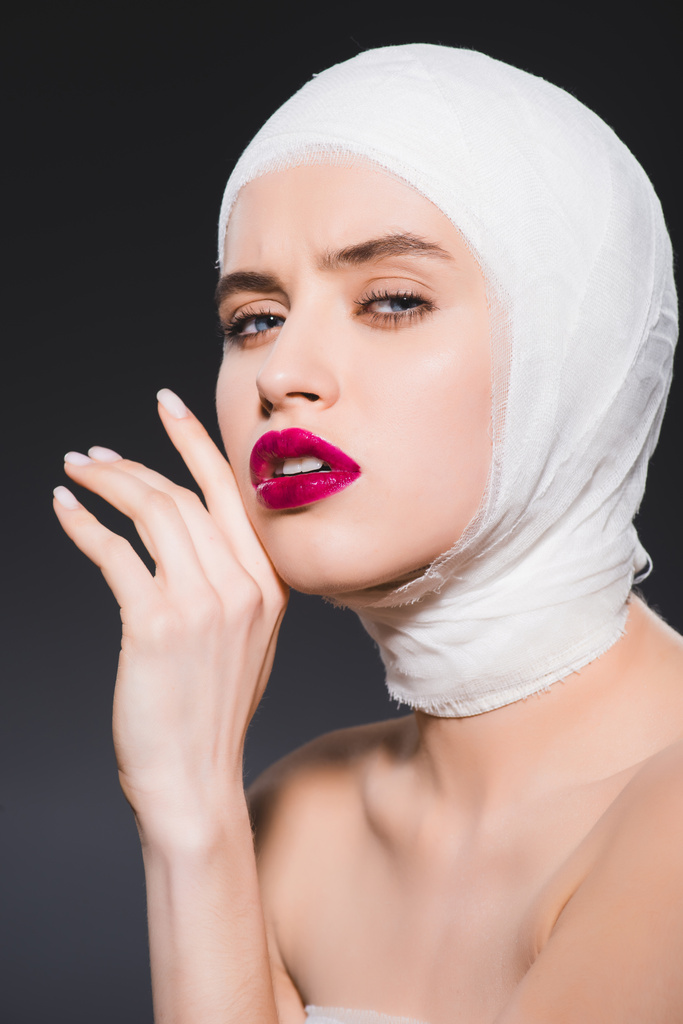young woman with bandaged head and crimson lips touching face isolated on grey  - Photo, Image