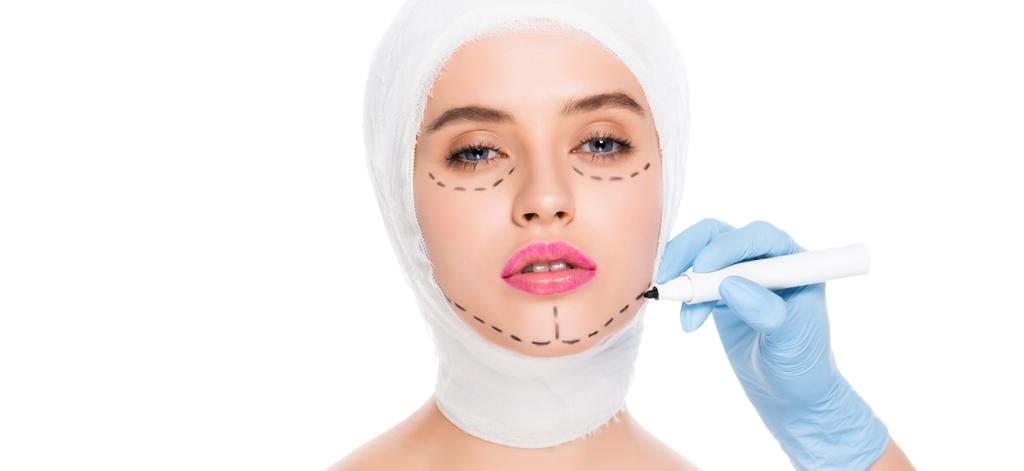 白い上に孤立した顔にマークを持つ女性の近くにマーカーペンを保持しているラテックス手袋のプラスチック外科医のパノラマ写真  - 写真・画像