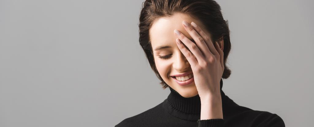 tiro panorâmico de mulher jovem alegre em preto jumper cobrindo olho isolado em cinza
  - Foto, Imagem