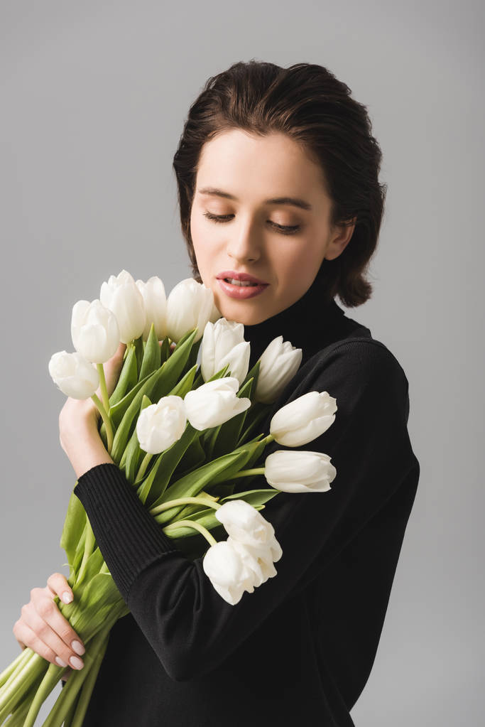 attraktive brünette Frau mit Blick auf weiße Tulpen isoliert auf grau  - Foto, Bild
