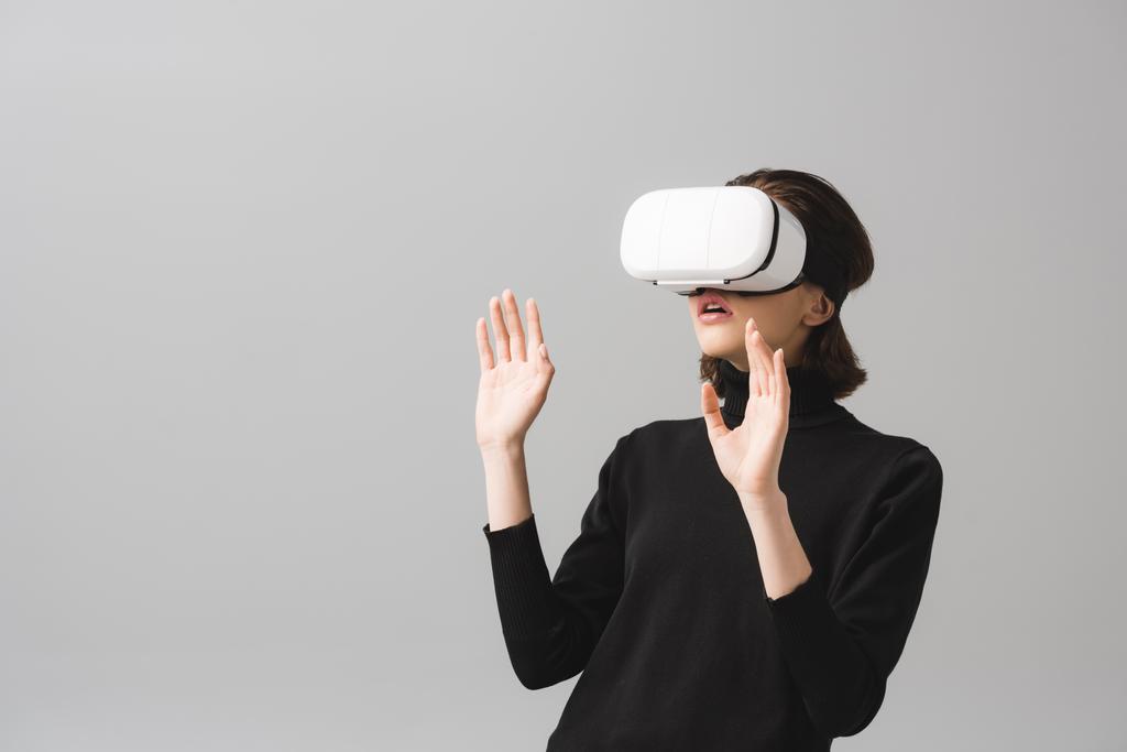 überraschte brünette Frau trägt Virtual-Reality-Headset, während sie isoliert auf grau gestikuliert  - Foto, Bild