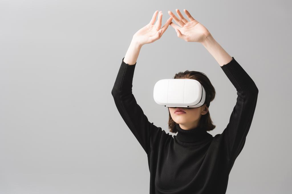 μελαχρινή γυναίκα φορώντας ακουστικά εικονικής πραγματικότητας, ενώ στέκεται με τα χέρια πάνω από το κεφάλι απομονωθεί σε γκρι  - Φωτογραφία, εικόνα