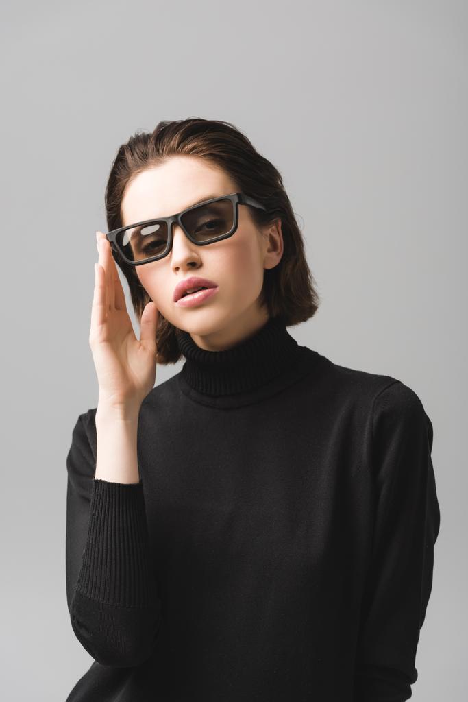 atraktivní mladá žena dotek slunečních brýlí izolovaných na šedé  - Fotografie, Obrázek