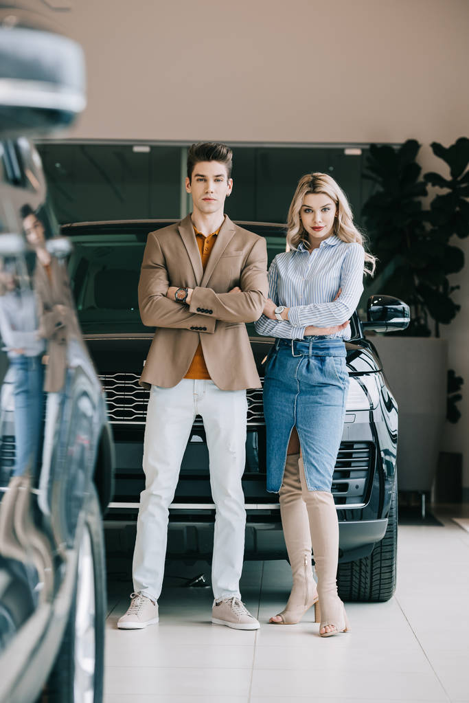 knappe man en mooie blonde meisje staande met gekruiste armen in de auto showroom  - Foto, afbeelding