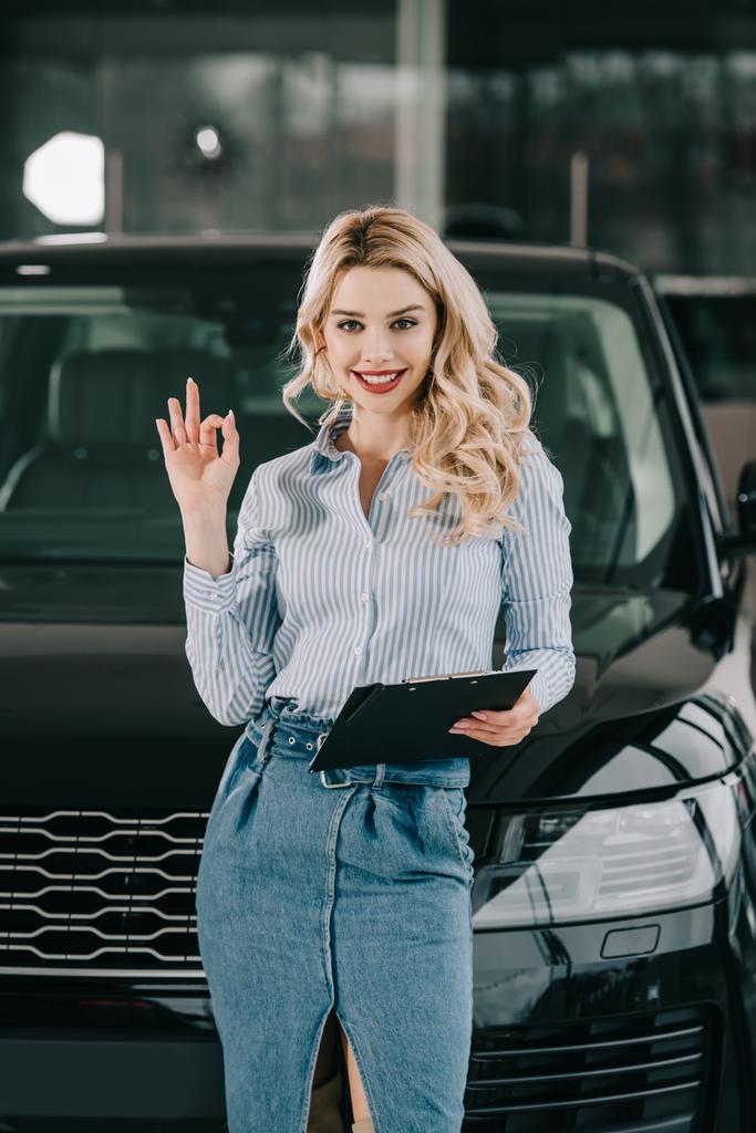 fröhliche blonde Autohändler hält Klemmbrett und zeigt ok Zeichen in Auto-Showroom  - Foto, Bild
