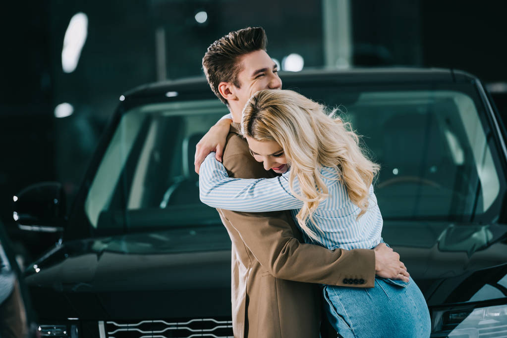 wesoły blond dziewczyna przytulanie szczęśliwy mężczyzna stojąc w pobliżu samochodu  - Zdjęcie, obraz
