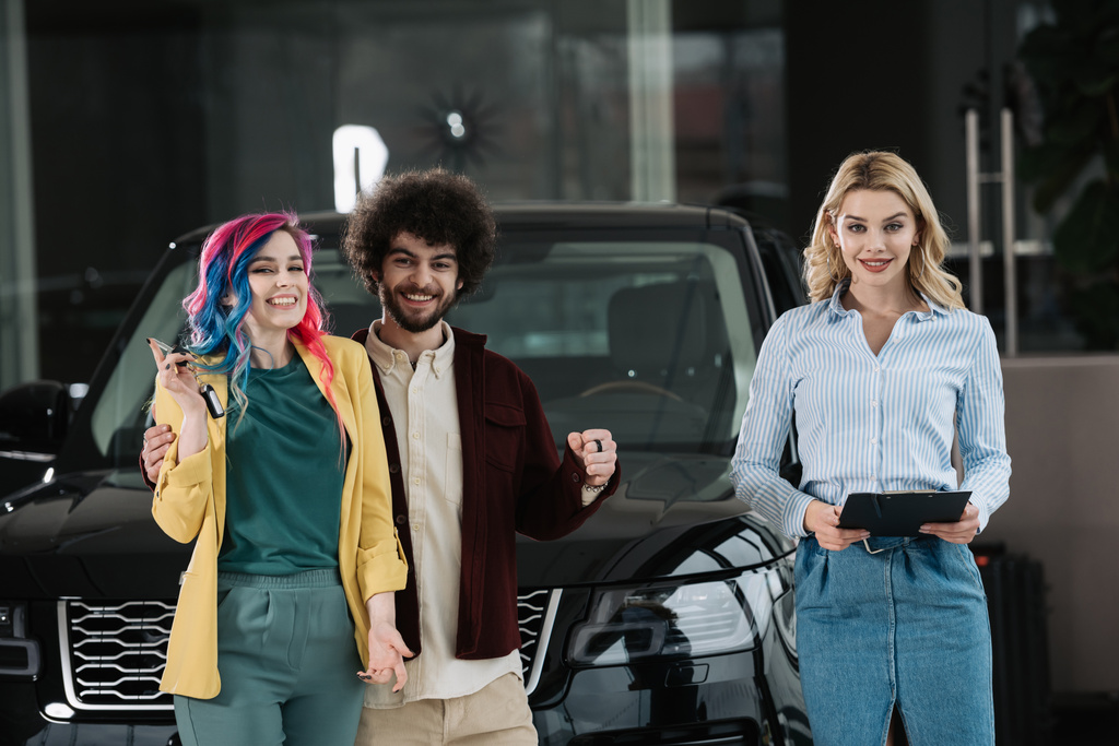 attractive blonde car dealer holding clipboard near cheerful man and woman  - Photo, Image