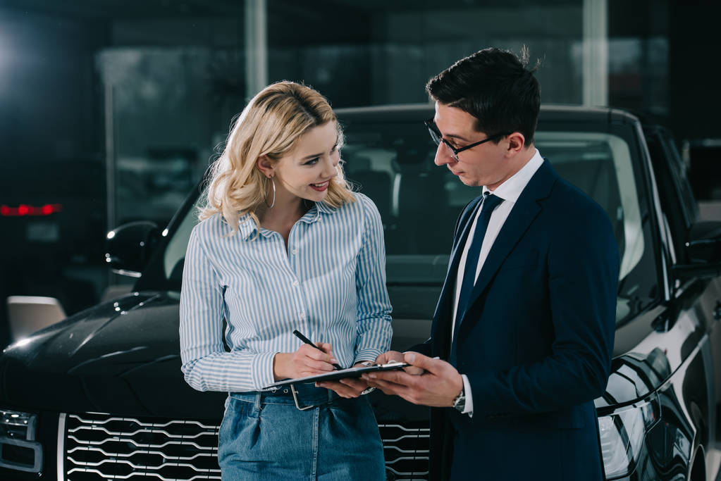 cheerful blonde woman signing contract and looking at car dealer  - Photo, Image