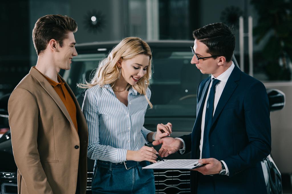 gelukkige man te kijken naar blonde vrouw staande in de buurt van auto dealer en ondertekening contract  - Foto, afbeelding