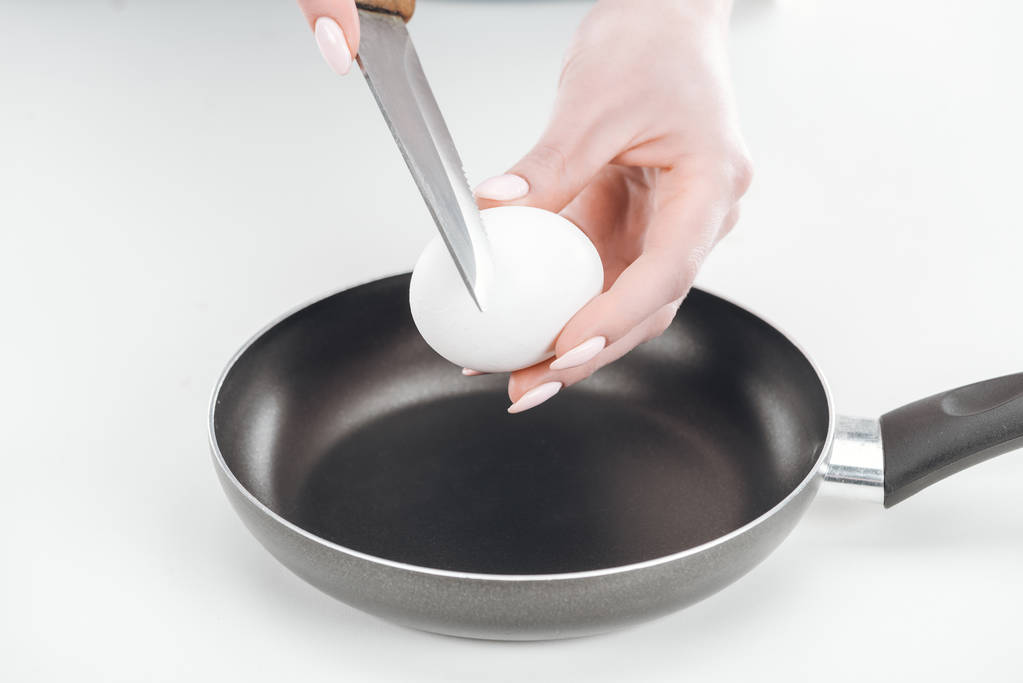 cropped view of woman holding chicken egg and knife near pan on white background - Photo, Image