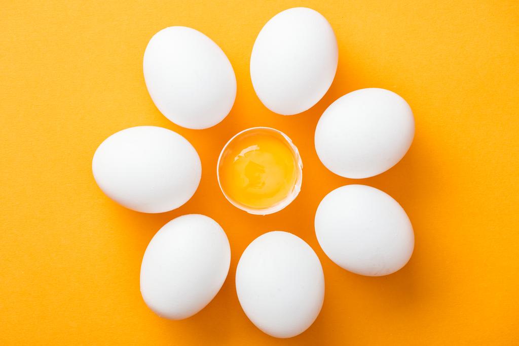 Widok z góry na całe białe jaja i rozbite jeden z żółtkiem na jasnym pomarańczowym tle  - Zdjęcie, obraz