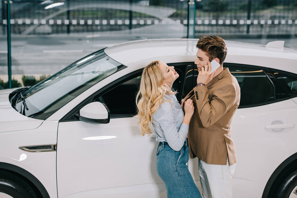 aantrekkelijke blonde vrouw aanraken jas van de mens te praten over smartphone in de buurt van auto   - Foto, afbeelding