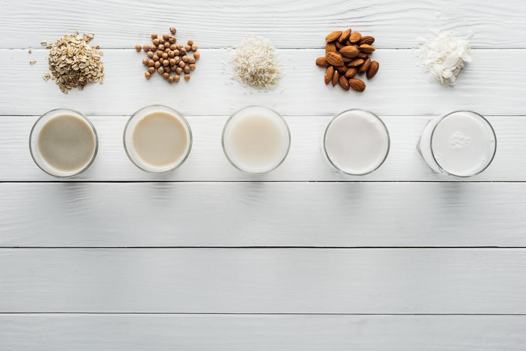 Κορυφαία θέα των γυαλιών με καρύδα, ρεβίθια, βρώμη, ρύζι και γάλα αμυγδάλου σε λευκή ξύλινη επιφάνεια με συστατικά - Φωτογραφία, εικόνα