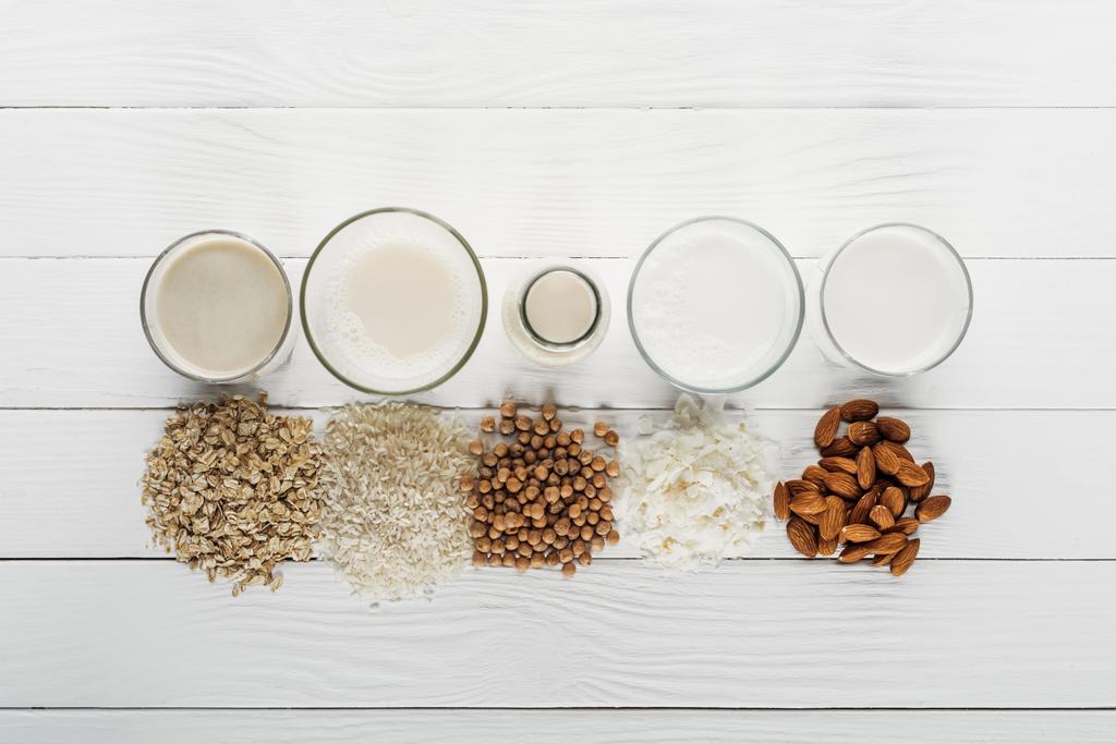 Κορυφαία όψη γυαλιών και μπουκαλιού με καρύδα, ρεβίθια, βρώμη, ρύζι και γάλα αμυγδάλου κοντά σε συστατικά  - Φωτογραφία, εικόνα