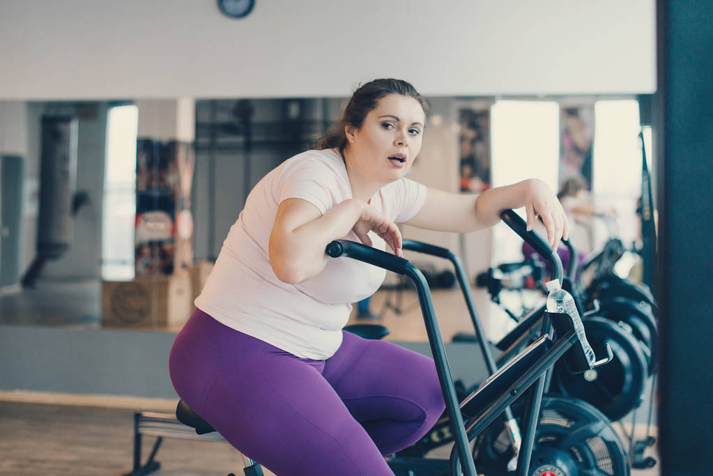 太った女性は、固定自転車に疲れて座る - 写真・画像