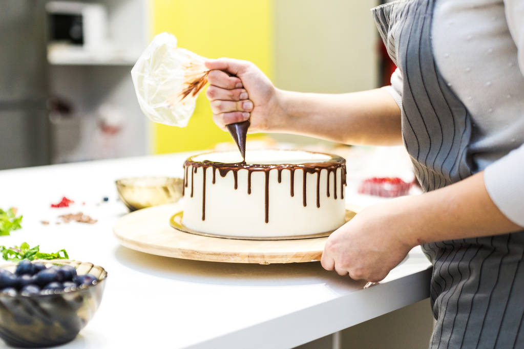 A cukrász összenyomja folyékony csokoládét egy cukrásztáskában rá egy fehér krém keksz torta egy fából készült állvány. A koncepció a házi készítésű péksütemények, főzősütemények. - Fotó, kép