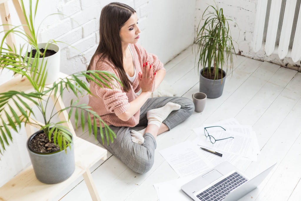 Young happy woman sits on the floor in a yoga pose in a bright apartment and works behind a laptop, a freelancer girl meditates while working. excitement and inspiration - Photo, Image