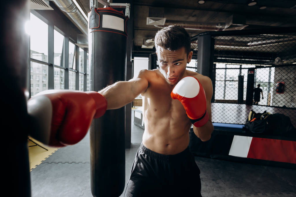Sportivo in rosso guanti da boxe con un torso nudo colpisce sacco da boxe in palestra sullo sfondo del ring di boxe
 - Foto, immagini