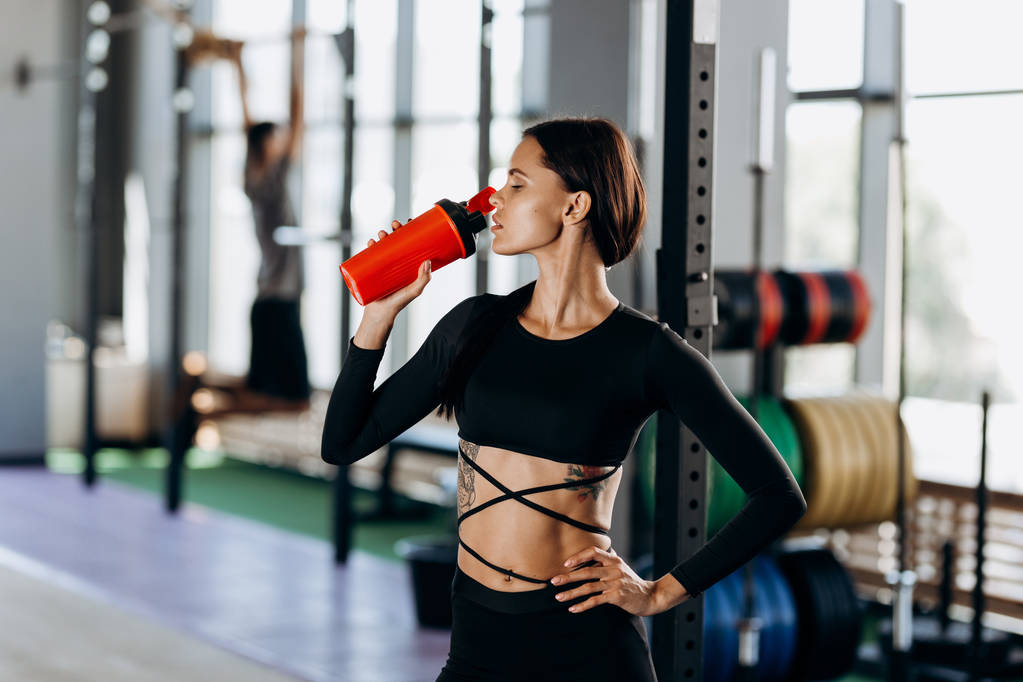 Menina de cabelos escuros magro vestido com sportswear preto bebe água no ginásio perto do equipamento desportivo
 - Foto, Imagem