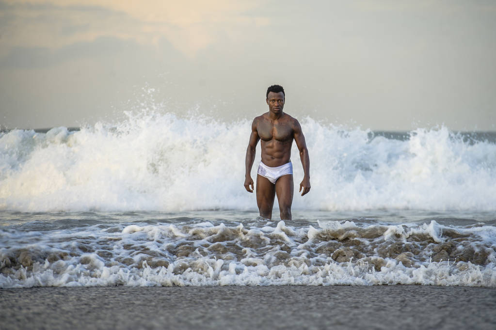 attractive and fit black African American man with sexy muscular body and sixpack abs posing cool on beach with big sea waves and foam around in beauty and health concept - Photo, Image