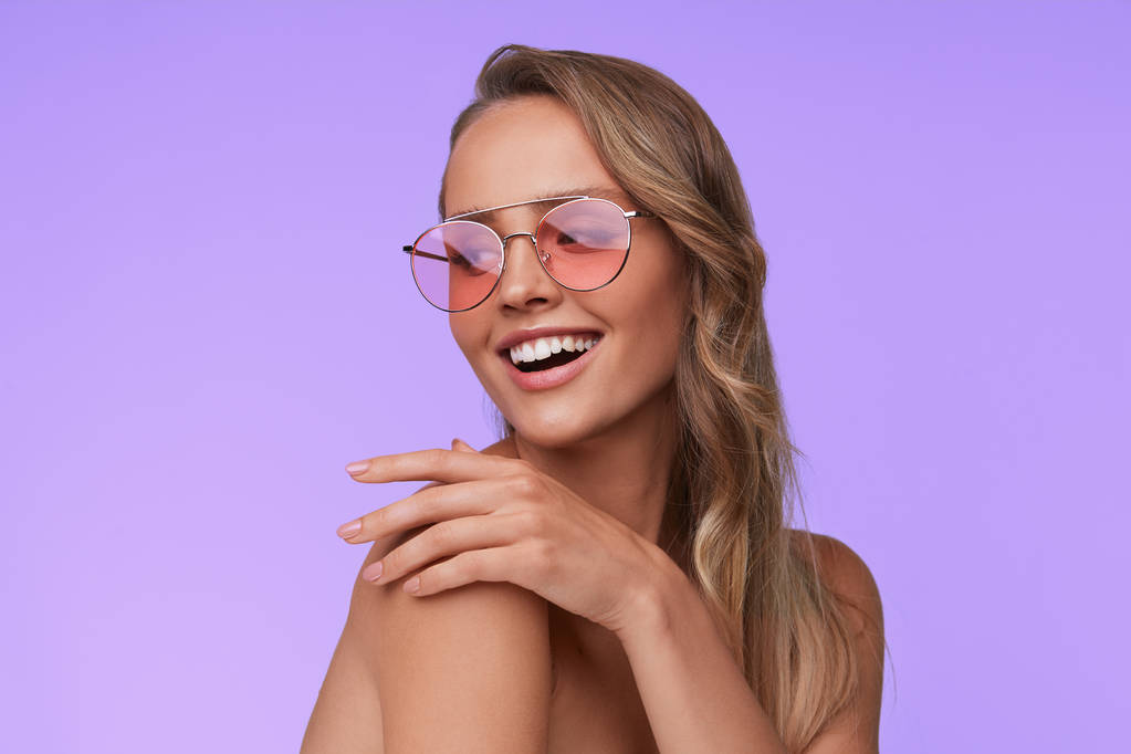 ピンクのサングラスを身に着けている完璧なメイクアップと美しい若い女性の肖像画。ライラックの背景にポーズのアビエイターサングラスでの笑顔のファッションモデル。スタジオショット。夏休み. - 写真・画像