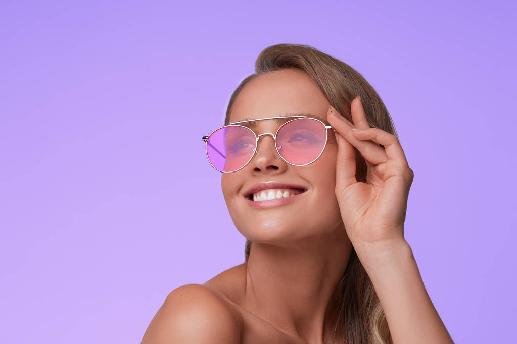 Portrait of beautiful young woman with perfect makeup wearing pink sunglasses. Smiling fashion model in aviator sunglasses posing on lilac background. Studio shot. Summer vacation. - Photo, Image