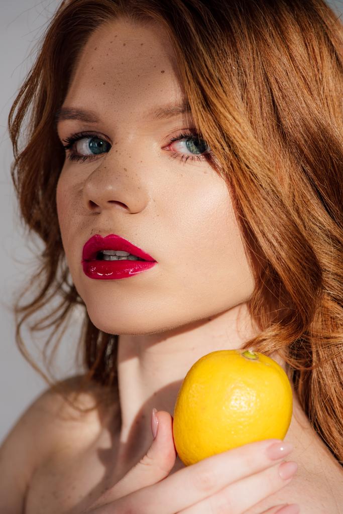 schöne rothaarige Mädchen mit roten Lippen hält Zitrone und posiert isoliert auf grau - Foto, Bild